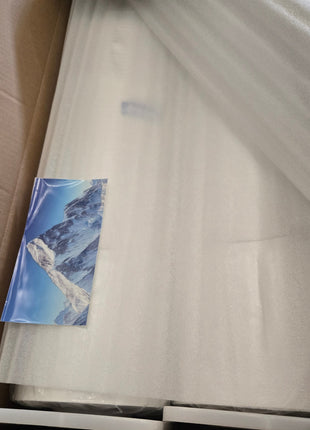 A large white rectangular item wrapped in protective foam and plastic inside a brown cardboard box, with a small printed card featuring a blue mountain scene tucked near the left side. A hand is visible at the bottom right.